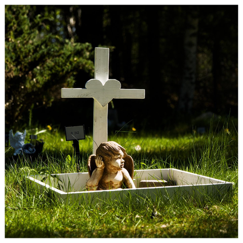 En fyrkantig bild på en grav för ett djur. På ett vitt kors sitter ett hjärta. Graven är omgiven av en vit rektangel och i den sitter en guldfärgad ängel. Grönt gräs runtomkring.