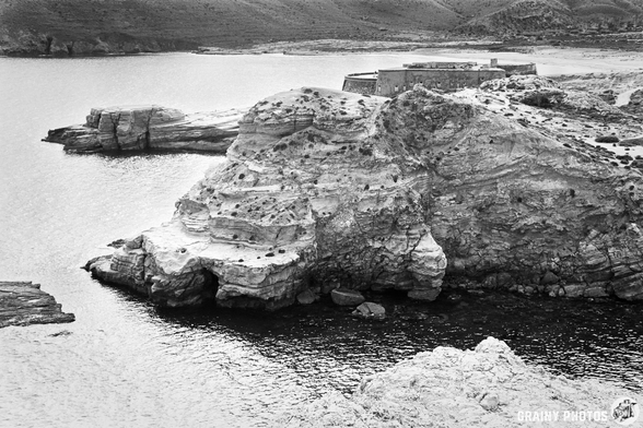 A black and white image of rocky cliffs by a calm sea, featuring a small fort at the top of the cliff, surrounded by rugged terrain and water reflecting the landscape.
