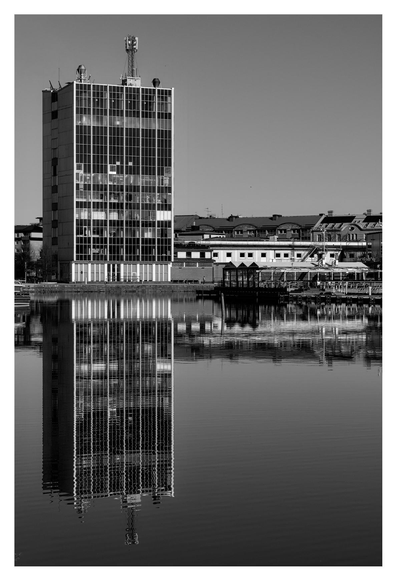 En stående svartvit bild på en hög, smal byggnad som speglas i vatten. Himmelen molnfri.