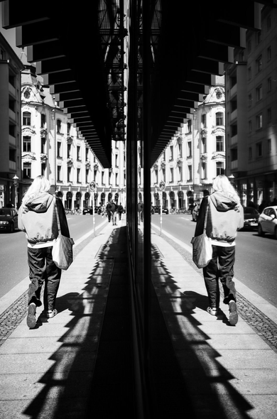 The image is a black and white photograph taken from a low angle, capturing a street scene with a striking play of reflections. The central focus is a large, highly reflective glass surface that bisects the image vertically. This reflection creates a mirrored, almost symmetrical composition.

On the left side of the image, in the "real" world, a person with long, blonde hair is walking away from the camera along a sidewalk. They are wearing a hooded jacket, dark pants, and carrying a light-colored tote bag over their shoulder. Their shadow, cast by the bright sun, stretches out in front of them, creating a sharp, angular pattern on the sidewalk due to the overhang of a modern architectural structure above. The sidewalk is paved with stone tiles and bordered by a curb and the road.