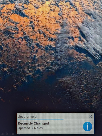 A digital notification window appearing over a desktop wallpaper. The title of the window is "cloud-drive-ui". The message reads "Recently Changed", followed by a status update: "Updated 356 files." A blue progress bar is visible near the top of the window. 

The wallpaper features a satellite-style view of a textured landscape. It displays earthy tones of orange and brown contrasted with dark blue and grey areas. 

This photo is a physical shot of a computer monitor, the individual pixels and a slight moiré pattern is visible.

This notification indicates that a cloud storage service has recently finished syncing a large batch of files to the local machine.