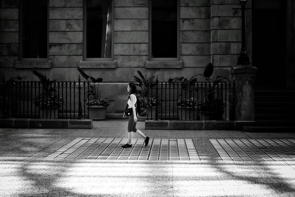 A woman walks past a large stone building with large rectangular windows and a steel fence in front of it. The woman wears a white top and a long dark skirt and she is walking on a square tiled surface with shadows crossing it. This is a black and white image.