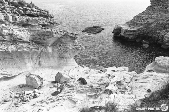A black and white landscape featuring rocky cliffs descending into a calm sea, with scattered boulders and sandy terrain in the foreground, capturing a tranquil coastal scene.