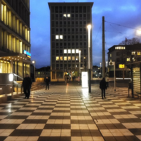 Städtische Straßenszene in der Morgendämmerung: seelenlose 60er-Jahre-Gebäude säumen einen breiten karierten Bürgersteig, auf dem Passanten wandeln.