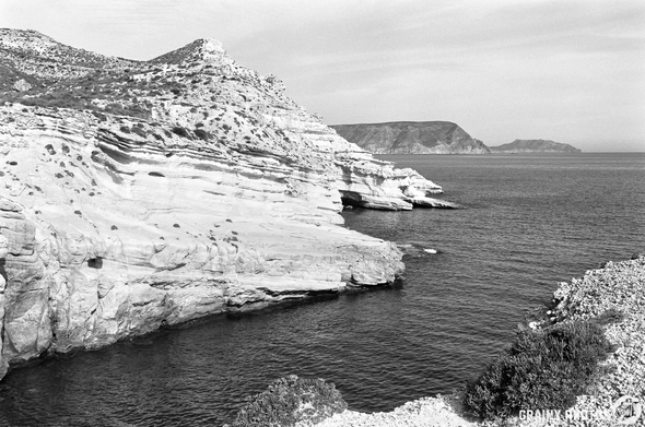 Black and white landscape featuring rugged cliffs and serene waters, with distant hills on the horizon. The rocky shoreline is accentuated by natural textures and vegetation, creating a tranquil coastal scene.