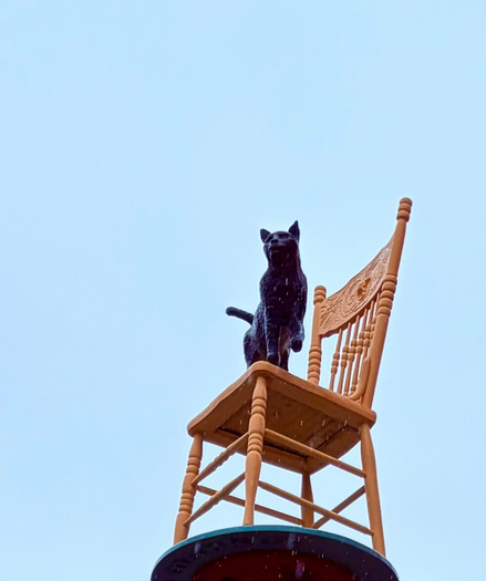 Orange kitchen chair on top of a pedestal. On the chair is a black cat sculpture. The cat was a resident of the neighbourhood, both named Kensington.