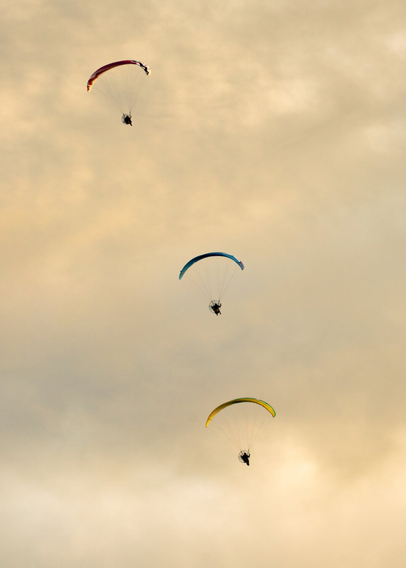 3 parachutes glide down through the morning sky, one underneath the other. The parachutes are red, blue, and yellow. The clouds have a golden hue.