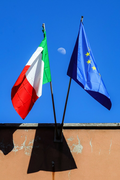 This is a photograph of two flags mounted on poles against a clear blue sky. The left flag is the Italian flag with green, white, and red horizontal stripes, and the right flag is the European Union flag which is blue with yellow stars. A partial moon is visible in the background between the two flags. The flags are mounted on a weathered, brown, textured wall, casting shadows. (The building is actually a school!)