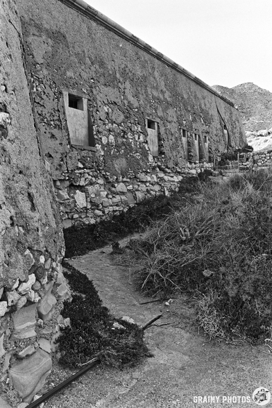 The rear of Castillo de San Ramón with partially closed windows, set against a backdrop of rocky terrain and sparse vegetation, captured in black and white. A narrow pathway leads along the structure, suggesting an abandoned or historical site.