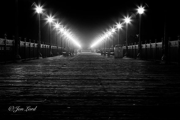 This is a night time, grungy, black and white photo in landscape format looking along a wooden pier lined with street lights that converge into the distance. The Embarcadero, San Francisco (2012).

The camera is about half a metre above the weathered and rough wooden planks that lay at right angles to the lens. The lighting above exaggerates the knurled and uneven texture of the well worn wooden surface. At the edges of the pier are two parallel rows of ornate, black metal railings. Spaced every three or four metres are dark metal street lights that are about three or so metres tall. Each street light is topped with a lantern that radiates outwards eight petal-like rays. Arranged along the edges are numerous park benches plus the odd large litter bin. Both the railings and street lights while in reality are parallel: in the image converge together with increasing distance. Above and to the sides of the scene is a pitch black and featureless sky. 

