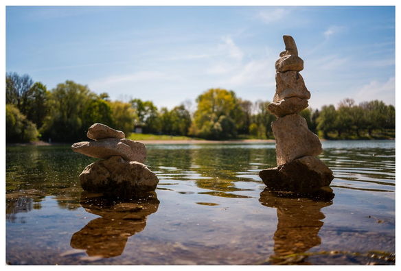 Zwei Türmchen aus hellen Steinen stehen in einem See. Im Hintergrund Bäume und Himmel.