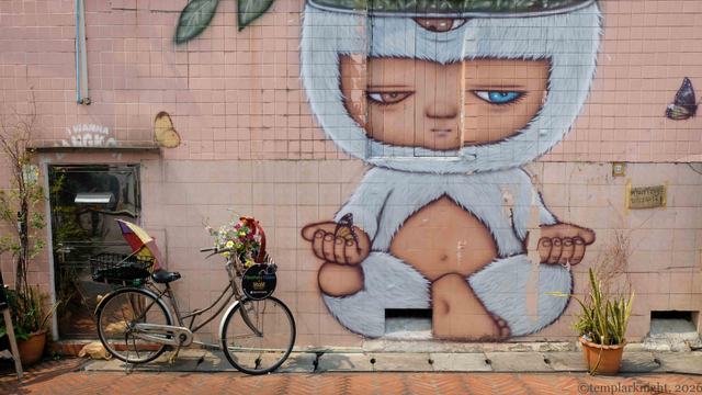 Color photo in landscape format of a large wall painting outside a restaurant on a sunny day. The painting is of a child-like boy sitting at a lotus position dressed in a white rabbit like suit. A woman bicycle parked outside the restaurant backdoor on the left 