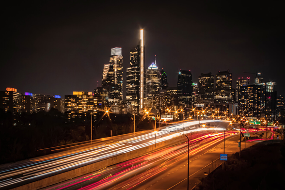 Philadelphia skyline at night with illuminated skyscrapers in the background and long-exposure light trails from cars streaming along a highway in the foreground.