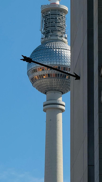 A close-up of the iconic television tower, the Berliner Fernsehturm, partially visible between two buildings against a clear blue sky. An arrow pointing towards the tower is visible on the right side.