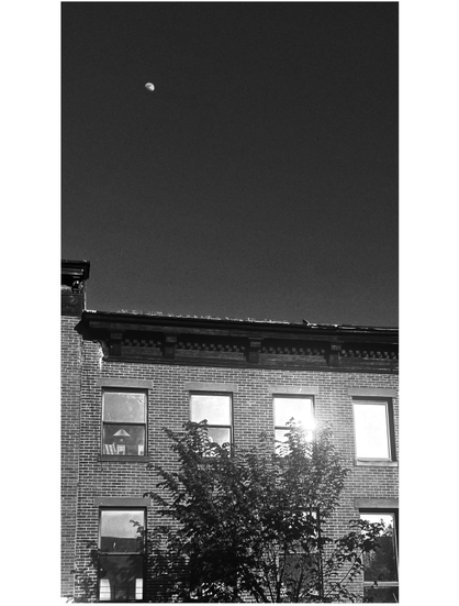 Black and white photo of a small waxing moon rising into a clear evening sky. Below is the facade of a brick apartment building, partially obscured by a slender tree. One of the building’s upper windows reflects the bright glare of the setting sun.