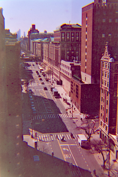 Looking down at a main road from the top of a building. Shadows of other buildings cast a jagged shape on the road. Several people were crossing that road