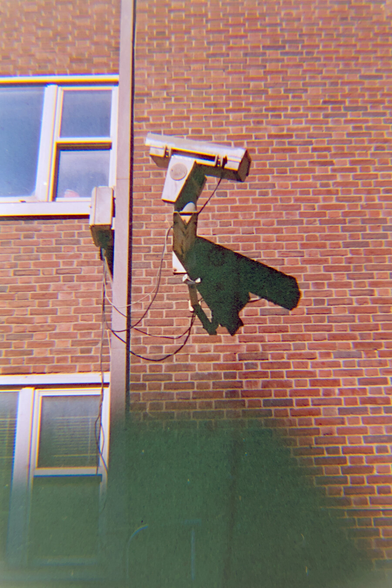 A surveillance camera on a brick wall casts a harsh shadow on the wall