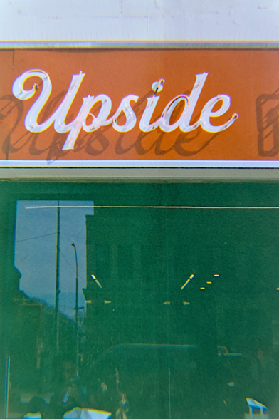 Red neonsign saying Upside in cursive is mounted in front of white letters of the same shape on red background. The sun casts a harsh shadow of the neon sign on the red background.