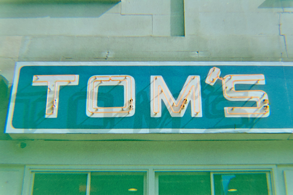 Red neon sign spelling Tom's in square font is mounted on white letters and blue background. The sun casts shadows of the neon signs on the blue background.