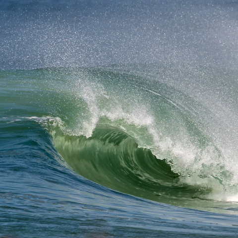 A colorful image of a barrelling wave breaking near the shore. The sea is mostly blue, and the wave is mostly green.
