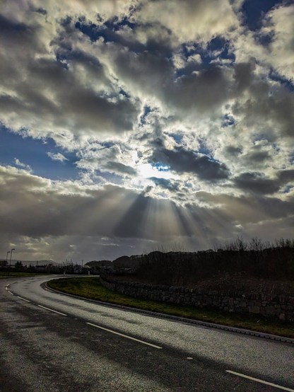 A winding asphalt road curves through a rural landscape under a dramatic sky where brilliant crepuscular rays of sunlight break through dark, heavy clouds.