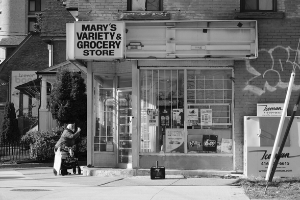 Looking across the street to a corner store. The sign reads Mary’s Variety and Grocery Store. Lottery signs line the lower edge of the barred windows. At far right, an ice machine sits against the brick wall. The edge of a graffiti tag can be seen on the wall. At far left, outside the store’s door, a woman leaning on a rollator gestures to someone in the store, unable to enter due to the concrete step.