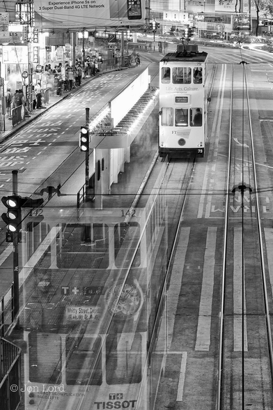 This is a long exposure, black and white, night time photo in portrait format of a busy city street with trams, cars and people blurred with their motion. Causeway Bay, Hong Kong (2014).

The camera is located on an overhead walkway about 3 or 4 metres above the street and looking between two parallel sets of tram (street car) tracks laid into the road. High above the street are two single power cables, one the power a trams on each track. Occupying the lower left corner and filling one quarter of the photo is a transparent, stationary, double deck tram, the road markings and rail tracks clearly visible through the tram. The tram is mainly white with an advert for Tissot watches and a band running between the two decks containing multiple images of their watches. On the back is a roller blind sign with the words 'Whitty Street Depot' and small Chinese characters below. A short distance ahead of our ghostly tram is a further tram, this one also white but has been stationary for a while, so is viewed as a solid object. To the left of the tracks is a covered tram stop with two traffic lights with all the lights lit. On the far left is the side of the street with a pavement, busy with a blurred crowd and some stationary people waiting for a buses. Behind are brilliant white lights of numerous shop fronts. Ahead of the second tram, the road turns 45º to the left and traffic can be seen on the far side of the street, coming towards the camera, again blurred. 