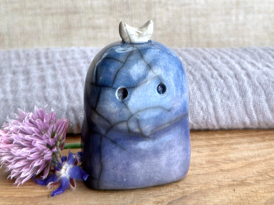 a kodama forest spirit kami raku sculpture glazed in a bottom-to-top gradient of indigo-violet to blue. it has a gentle face, and a white crescent moon on its head. it looks somewhat concerned. it's standing on a piece of chestnut wood with grey muslin and natural linen behind. next to it on the left are a blue borage flower and a purple chive flower with a green stem.