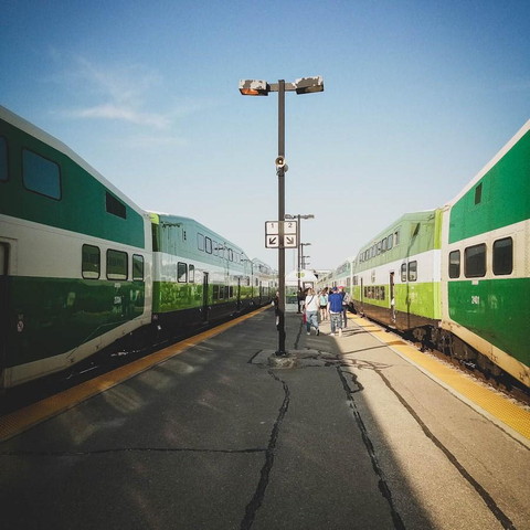 While standing in the middle a outdoor platform of a train station, green and white trains can be seen on either side. People can be seen walking and a tall light pole is also seen. Its is a sunny clear spring day with few soft clouds in the sky. 
