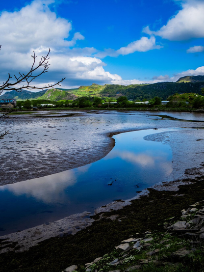 A scenic view of Llyn Bach (little lake) in Porthmadog at low tide. A channel winds through tidal mudflats reflecting the blue cloudy sky. In the background green mountains stand in the sun.