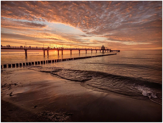 A scenic sunset view over calm waters, featuring a long pier with silhouetted figures and warm hues of orange and purple in the sky. Gentle waves lap at the shore in the foreground.
