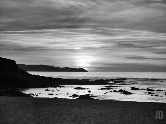La imagen muestra un paisaje costero en blanco y negro, con un atardecer sobre el mar. En primer plano se ve la orilla rocosa con pequeñas piedras y charcos de agua, mientras que al fondo se distinguen siluetas de colinas y un cielo cubierto de nubes finas que reflejan la luz del sol en el horizonte. La escena transmite tranquilidad y una sensación de calma natural.