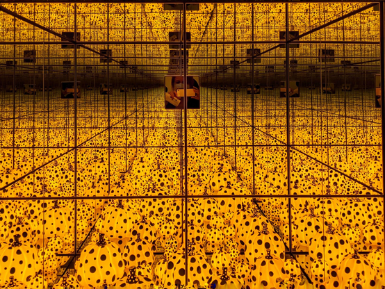 Ein verspiegelter Raum, gefüllt mit scheinbar unzähligen schwarz gepunkteten gelben Kürbissen. Im oberen Bereich ist eine kleine Schauluke zu sehen, durch die ich mit meinem Smartphone das Motiv, einen Infinity Mirror Room, fotografiere.