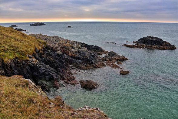 A rugged coastline at Ynys Llanddwyn, featuring jagged, dark rocky outcrops and patches of grassy vegetation. The turquoise waters gently lap against the shore, while small rocky islets dot the horizon under a soft, pastel-hued sky at dusk.