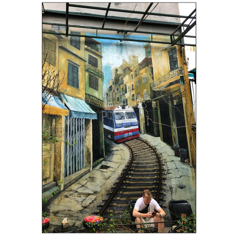 A train barrels down a narrow village road towards an unsuspecting white man sitting in a chair in the middle of the tracks. Actually, this is a benign scene inside Hidden Gem Cafe in Hanoi, Vietnam: the man is sitting against a wall on which the train is painted. The realistic mural depicts a tourist destination in Hanoi, Train Street, where a train regularly makes its way through a narrow passage between crammed houses that seem dangerously close to the tracks.
