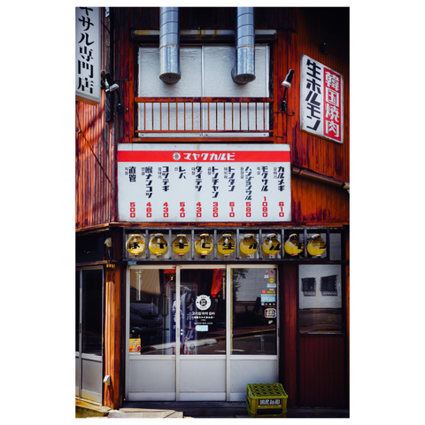 A frontal view of a small restaurant with a wooden exterior. A large menu board with Japanese text and prices is mounted above the entrance, and a row of yellow paper lanterns hangs just below it. Two metal exhaust pipes extend outward above a frosted window, and sliding glass doors form the entrance at street level. A green crate sits on the ground near the doorway.