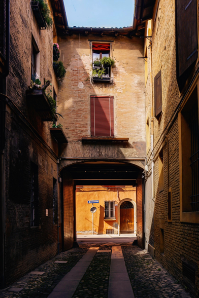 A photograph of a narrow, cobblestoney alleyway in Bologna. The alley has tall, aged brick buildings with wooden shutters and flower boxes. At then end there is an arched entrance/exit through which is a sunlit street with a yellow-orange building and a street sign. The light casts warmly on the textured bricks and pathway.
