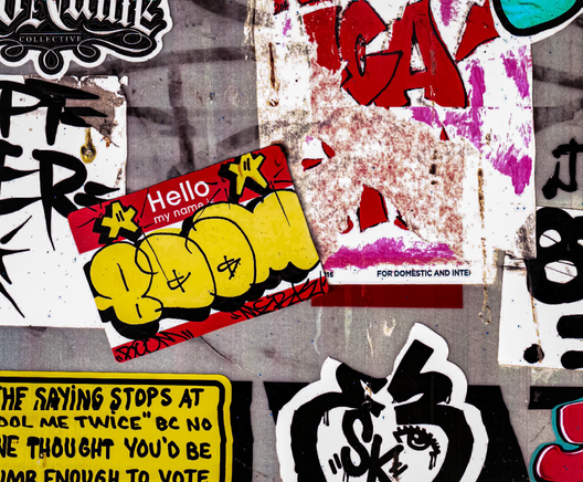 Close-up of a worn urban surface covered in overlapping stickers and graffiti tags, including a red “Hello my name is” label with a yellow bubble tag, peeling paper, and layered textures of paint, dirt, and adhesive residue.