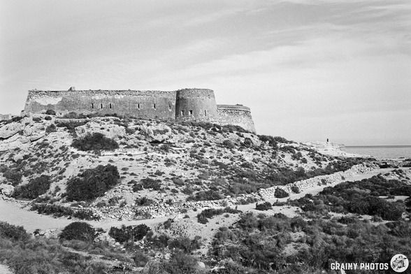 Castillo de San Ramón sits atop a hill, surrounded by rugged terrain and sparse vegetation, overlooking the sea under a cloudy sky. The scene captures the blend of natural beauty and architectural heritage.