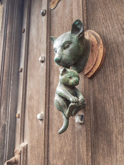 A photograph of a bronze door knocker on a weathered wooden door. The knocker is shaped like a cat's head holding a smaller, curled-up kitten in its mouth by the scruff.