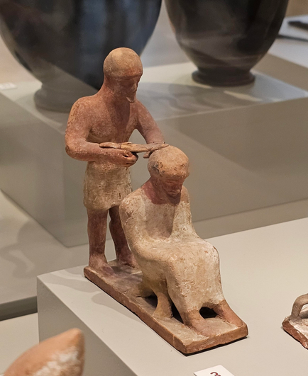 A terracotta figurine of a standing man cutting the hair of another man who is seated in front of him, shown on display in a museum setting 
