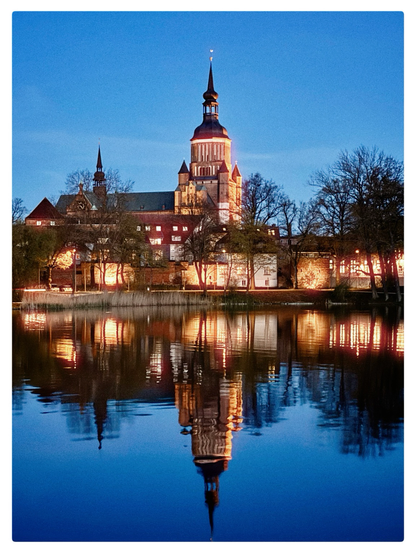A serene evening scene featuring a beautifully illuminated Gothic-style cathedral with a tall spire, reflected in a calm body of water. The building is surrounded by trees with soft, warm lights, creating a peaceful and picturesque atmosphere against the deep blue sky.