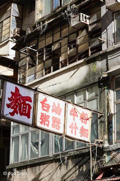 This is an urban colour photo in portrait format of the front of a decaying city building with an overhead street sign in Chinese characters. North Point, Hong Kong (2014).

The camera is angled upwards and at an angle of 45º to the front of this old, low-rise urban building. In view are the first, second and part of the third floors. The facade is heavily weathered with mould, rot and flaking paint. It has the beginnings of an urban botanical garden with various bushes, plants and moss growing from the many cracks and crevices. Extending outwards from the first floor and over the street is a rusting metal sign with three white squares. The left square has one large red Chinese character, while the middle and right have four characters arranged in a square within the white square. The first floor has large vertical rectangular aluminium window frames with white curtains behind, some torn and frayed. The second floor has older, small, horizontal rectangular galvanised metal frames, most rusting, all the windows caked in dirt, many of the windows broken. In the top right corner of that long since abandoned apartment or office is a corroding external air conditioning unit. Above is a small corrugated overhang with small bushes growing and hanging downwards. The floor above has large, rectangular windows, all caked in grime, some open. 