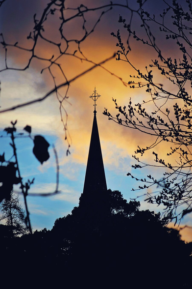 A tall, dark silhouette of a church spire dominates the centre of the frame. The spire is topped with an ornate iron cross. The background is a dramatic sunset sky with bands of deep orange, soft yellow, and pale blue clouds. Bare, dark tree branches frame the top and sides of the image, creating a natural border. The foreground shows the dark outlines of treetops. The overall mood is quiet and contemplative.