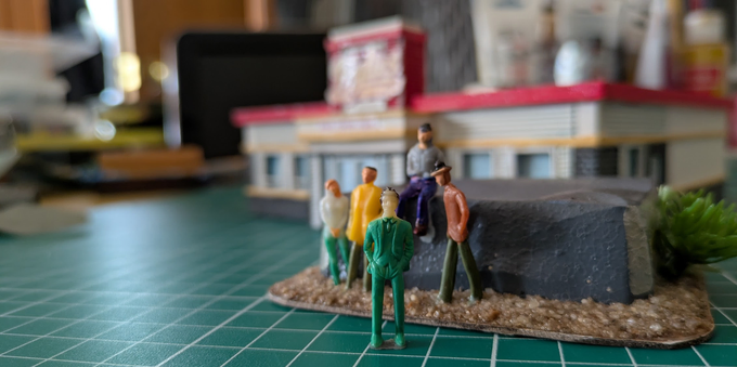 A group of five miniature plastic characters stand as if posing for the camera. Any distinguishing facial features have been removed. In the background, out of focus, a model of a fast food building looms.