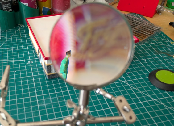 A small, faceless plastic diorama figure with black hair and green clothing is sharp underneath a magnifying glass. A distorted, blurred, out-of-focus model of a Frylies restaurant sits behind them. 