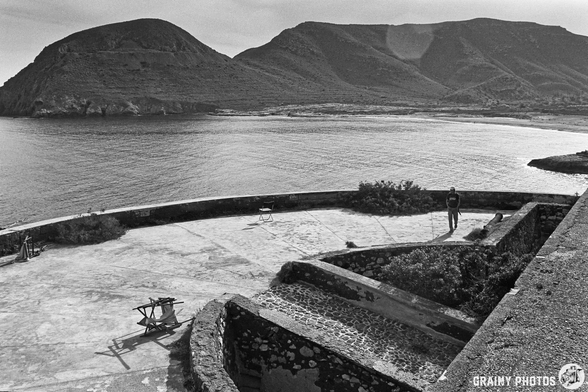 A serene black and white landscape featuring a coastal view from Castillo de San Ramón with rugged mountains in the background, a person standing on a stone terrace, and scattered outdoor furniture, evoking a sense of solitude and natural beauty.