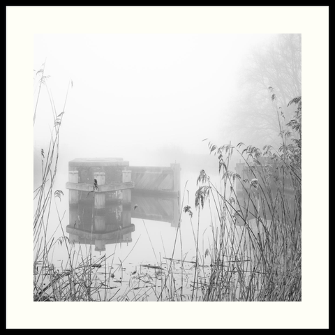b&w - Square format, high key photograph of part of a sluice gate along the Delftse Schie. Tall reeds stand in the fog-free foreground. In the middle ground, most of the gate is visible, lightly shrouded in mist. The background is almost entirely obscured by the early morning fog.