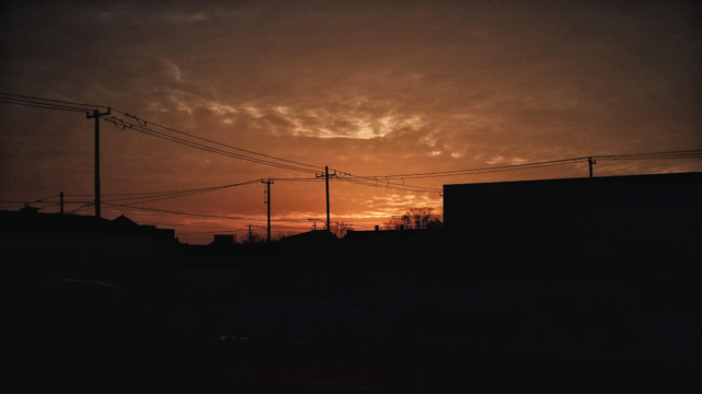 An orange sky is seen as the sun dips below the horizon. The silhouette of a building and utility poles and wires can be seen along the bottom. Some clouds are shown in the sky. 