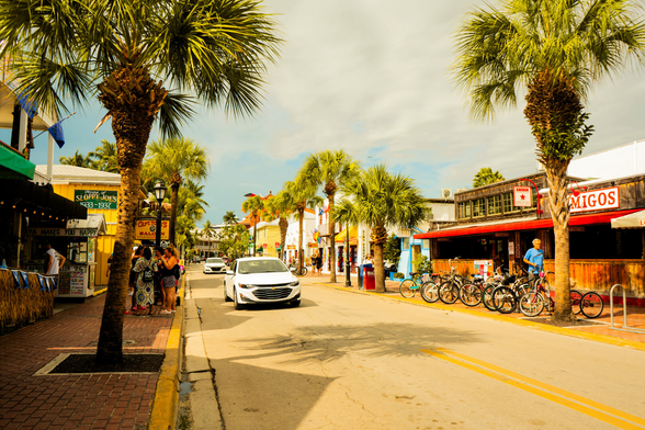 A group stands outside a line of bars on one side of the street, and palm trees divide the scene, with a runner on the other side. Two white compact cars drive down the middle.